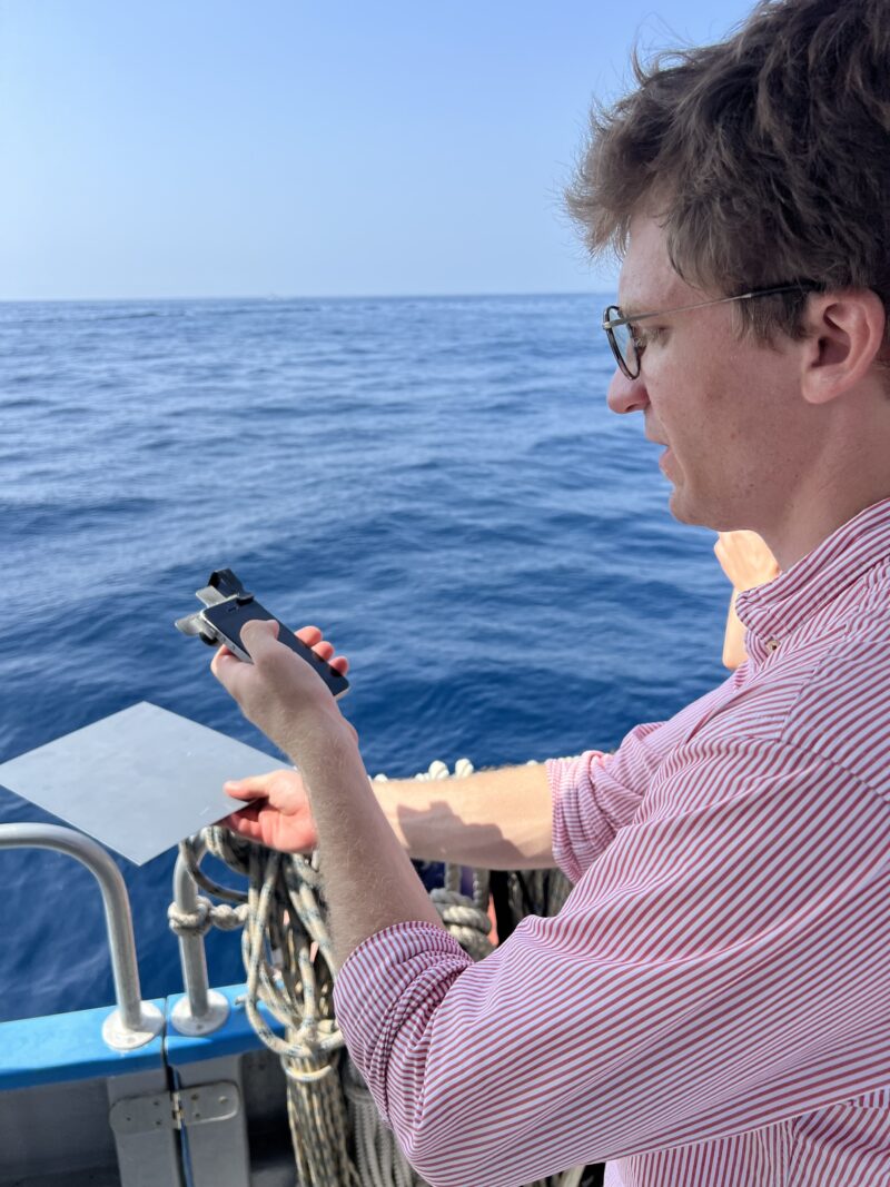 A researcher uses iSPEX2 with a smartphone and grey reference card above the sea